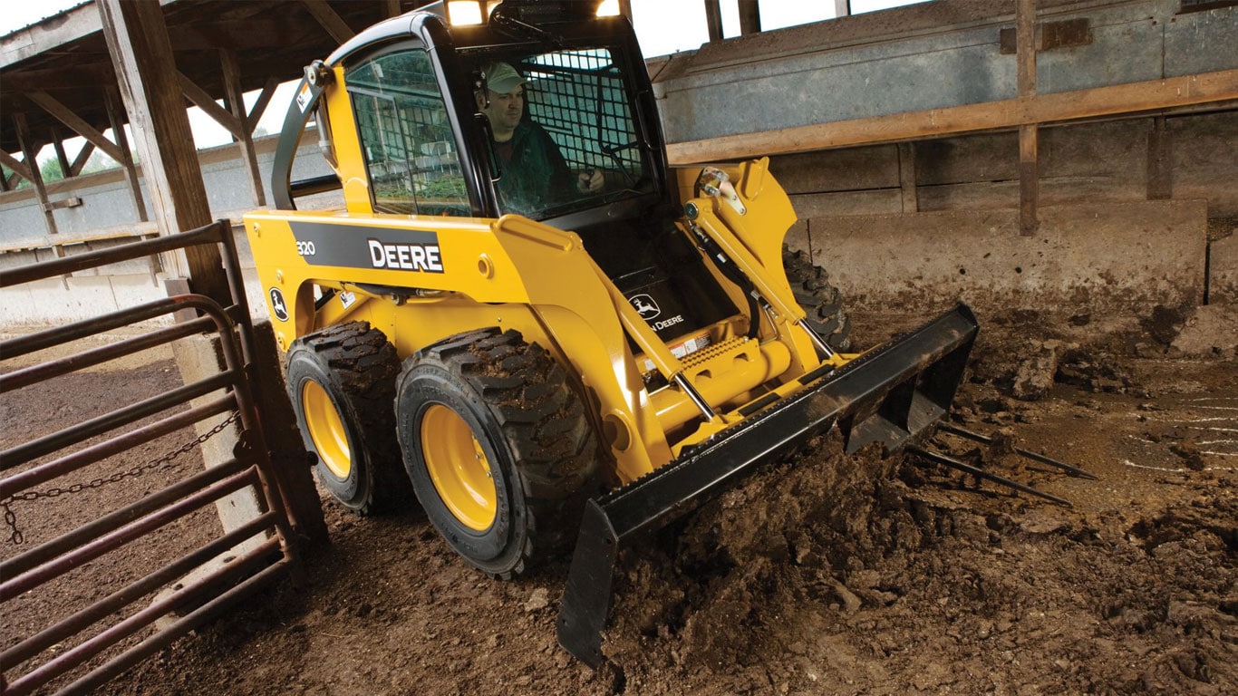 Une chargeuse à direction différentielle John Deere équipée de l’accessoire râteau dessoucheur-déracineur travaille sur une ferme.